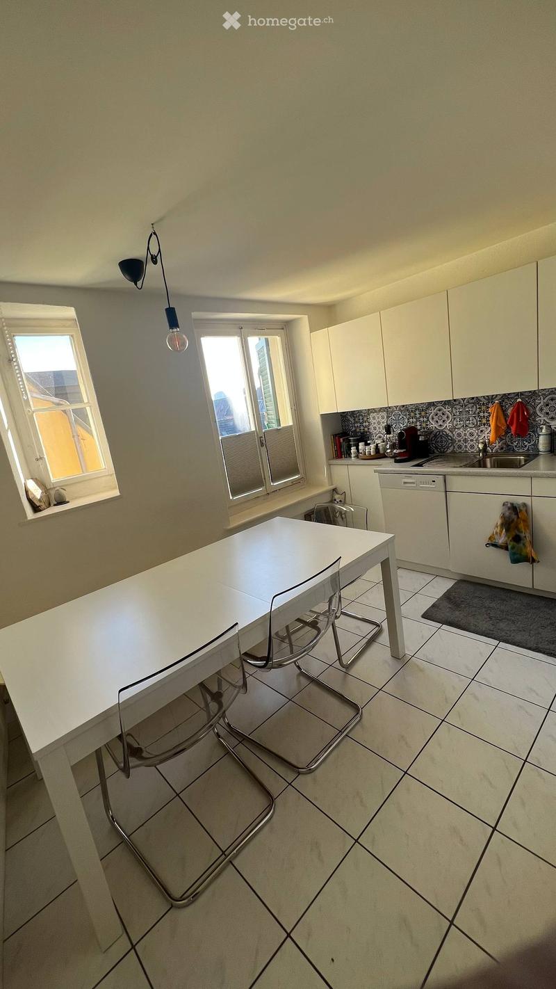 Modern kitchen with white cabinets, a white dining table with two chairs, a dishwasher, and a sink. The room features tiled flooring and large windows letting in natural light.