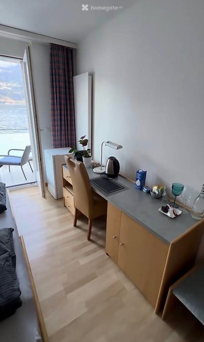 A furnished room with a desk, chair, lamp, and a small table with a cup and bottle. The room has wooden flooring and a view of the ocean through a large window.
