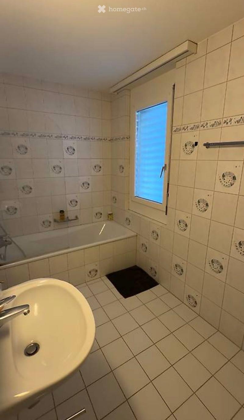 Bathroom with beige tiled walls and floor, a white sink with a chrome faucet, a bathtub, a mirror, and a towel rack. A small window with frosted glass is visible above the bathtub.
