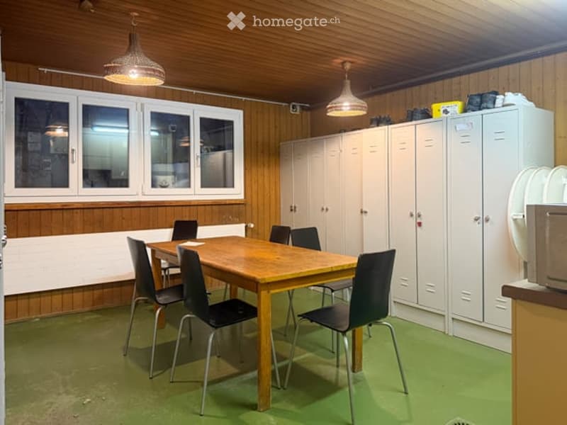 A room with wooden walls and ceiling, green flooring, a wooden table surrounded by black chairs, white lockers, and a microwave on a counter.