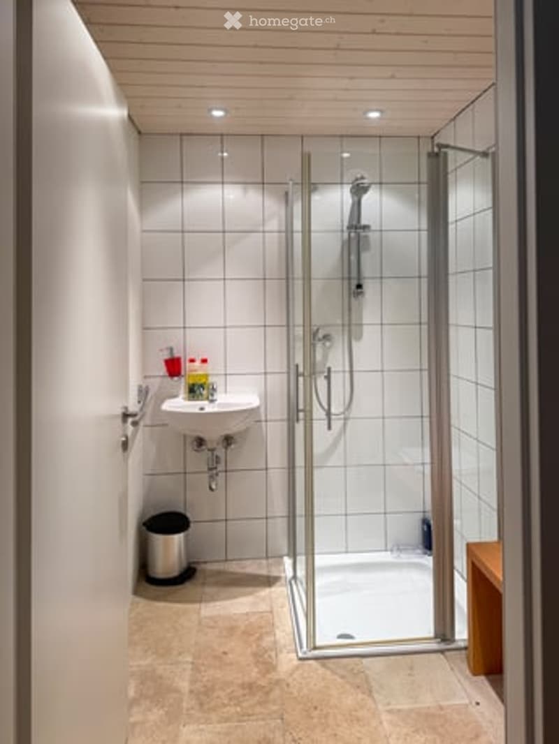 Modern bathroom with white tiled walls, a glass-enclosed shower, a sink with a faucet, and a small trash can.