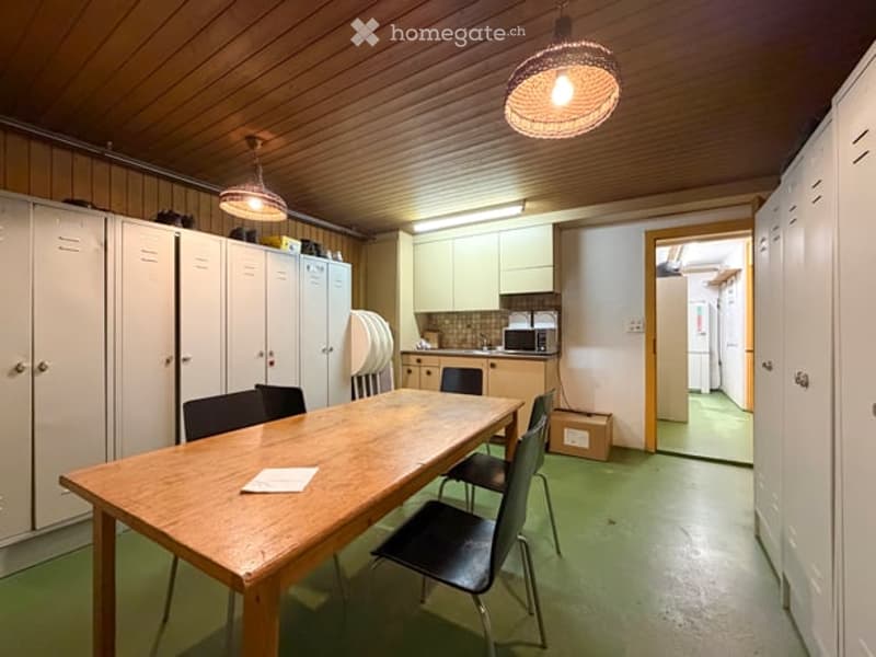 A room with a wooden table and chairs, white cabinets, and a small kitchen area with a microwave and refrigerator.