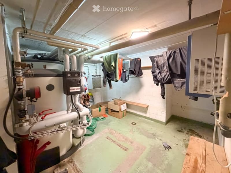 Unfurnished basement space with exposed pipes, a water heater, and storage boxes. Clothes are hanging on a rack, and the floor is concrete.