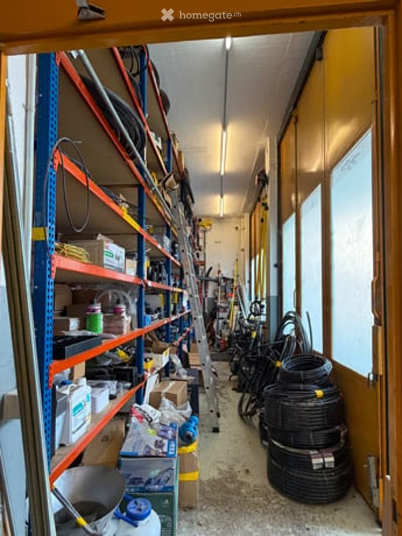 Storage area with metal shelving units filled with tools, boxes, and supplies. A ladder is positioned against the wall, and coiled black pipes are stacked on the floor.