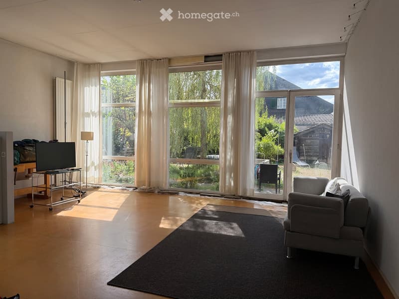 Bright living room with large windows and sliding glass doors leading to a garden, featuring a grey sofa, a coffee table, a TV, and a floor lamp.