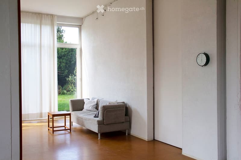 Bright living room with a grey sofa, wooden stool, and a wall-mounted clock. Large window with white curtains offers a view of a green garden.
