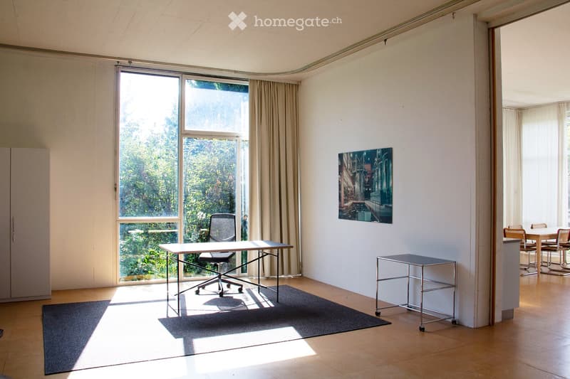 Bright, minimalist room with large windows, wooden flooring, and modern furniture including a desk, office chair, small table, and dining area with chairs and table.