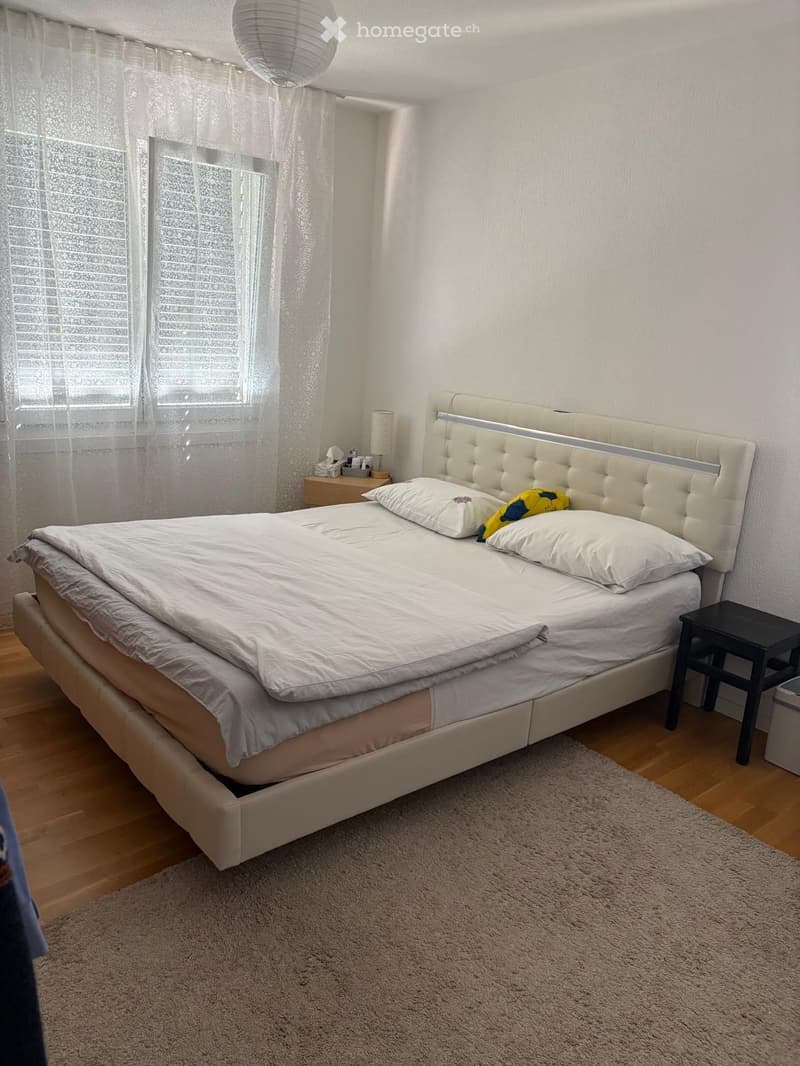 Modern bedroom with a large bed, white bedding, a tufted headboard, nightstand, and a small black stool.