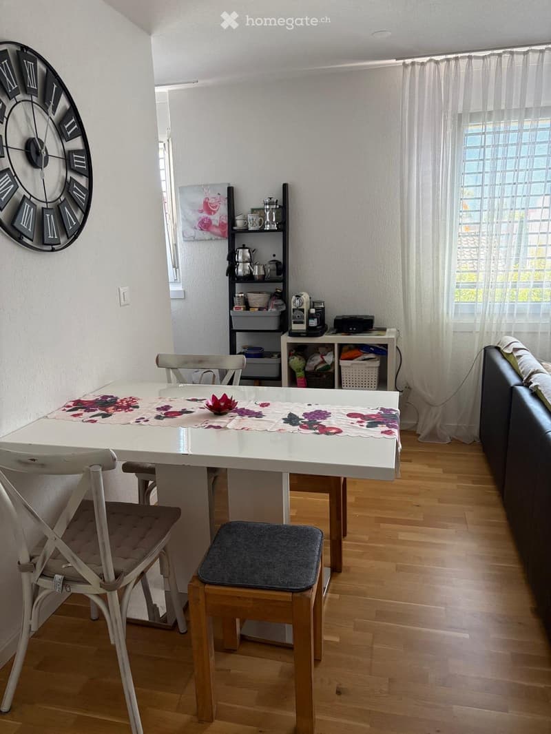 A modern dining area with a white table, floral placemats, and four chairs, including a wooden chair with a cushion. A sofa is visible to the right, and a small shelf holds kitchen items like a teapot, cups, and a coffee maker. A large wall clock and a fr