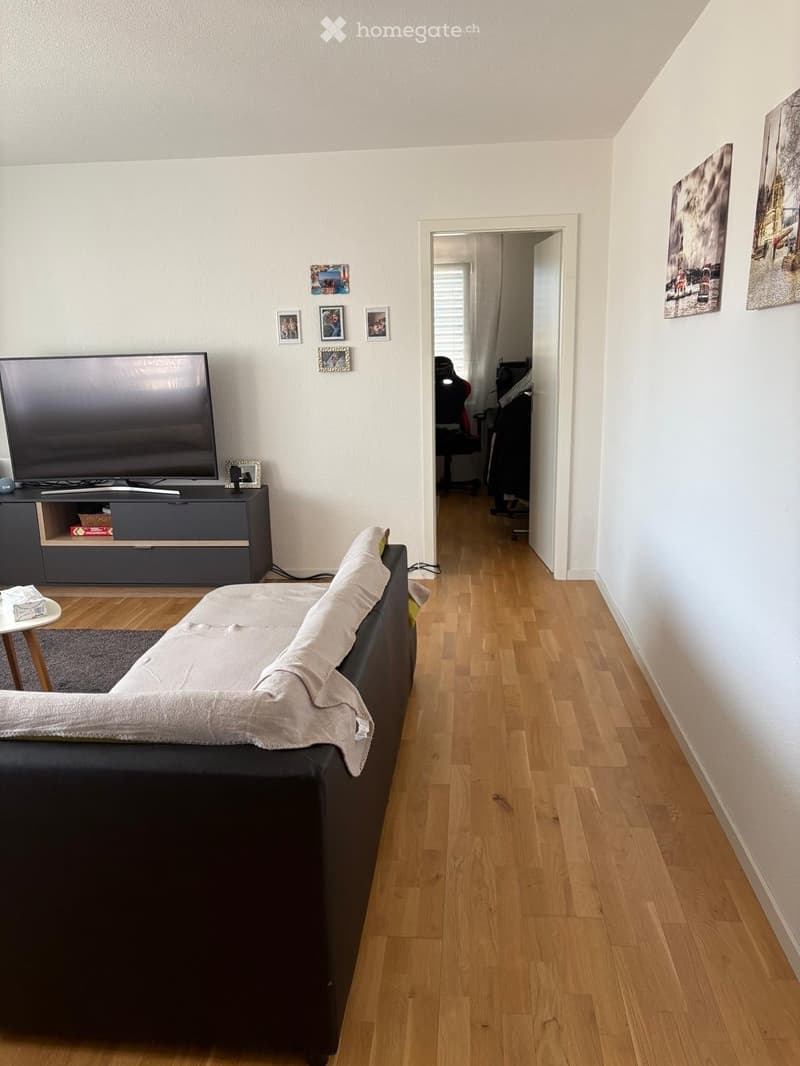 Living room with a grey sofa, a small table, a TV, and a chair in the background. The room has wooden flooring and white walls.