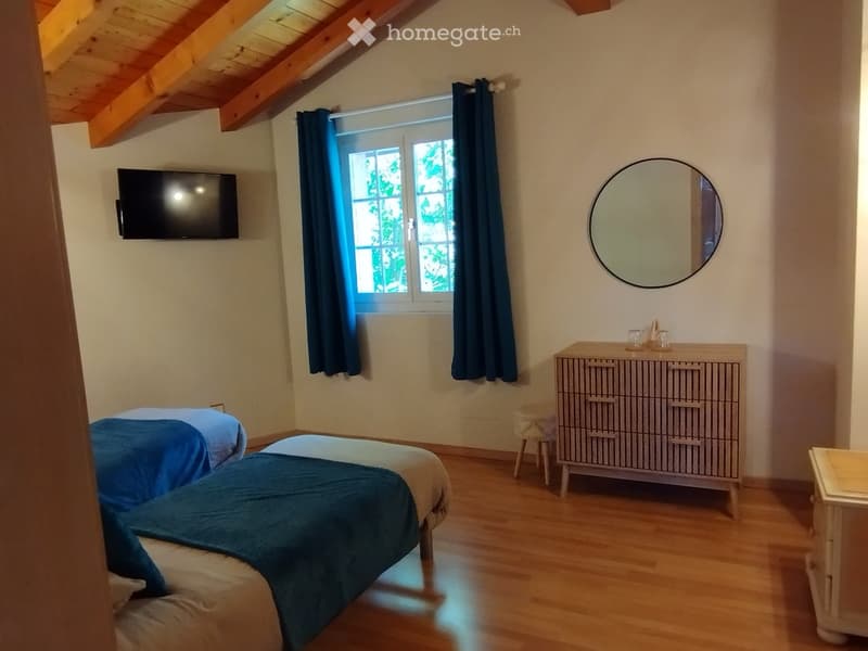 Cozy bedroom with wooden floors, two single beds, a dresser, a round mirror, and a television mounted on the wall.