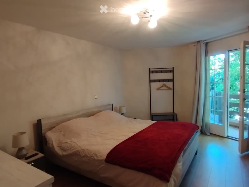 Cozy bedroom with a large bed, red blanket, nightstands with lamps, and a clothes hanger near the sliding glass door leading to a balcony.