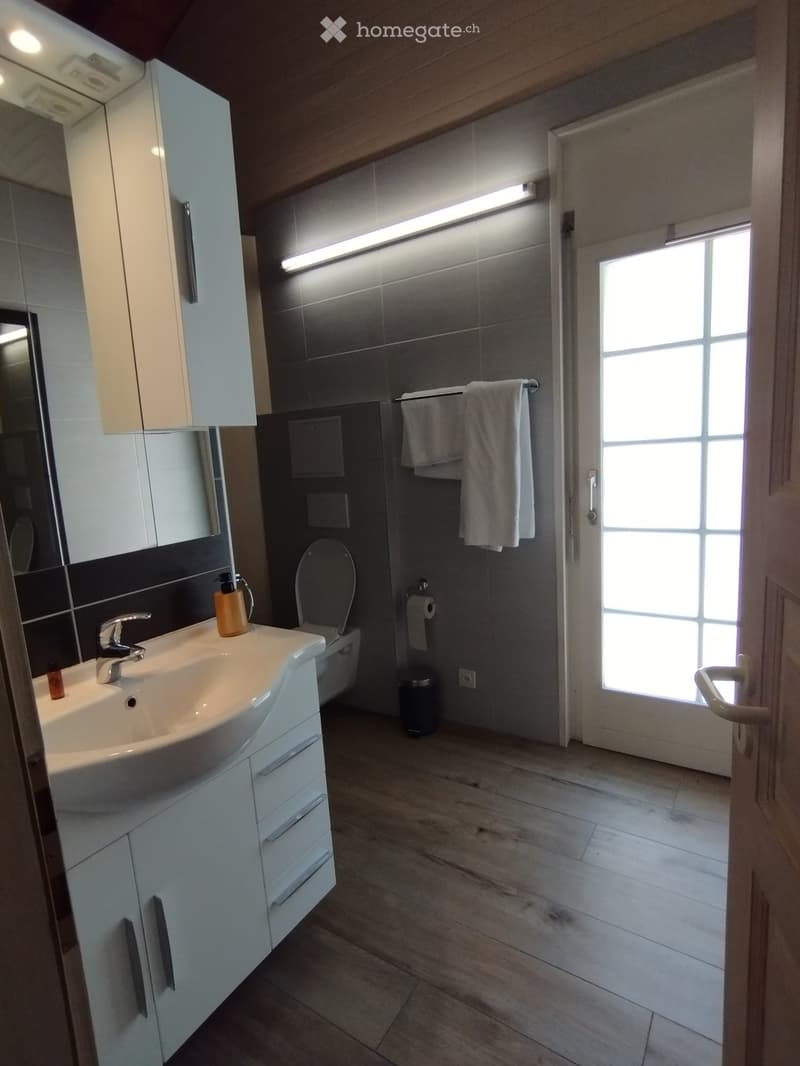 Modern bathroom with white cabinetry, a sink, a toilet, a mirror, and a towel hanging on a rack. The floor is tiled, and there is a window providing natural light.