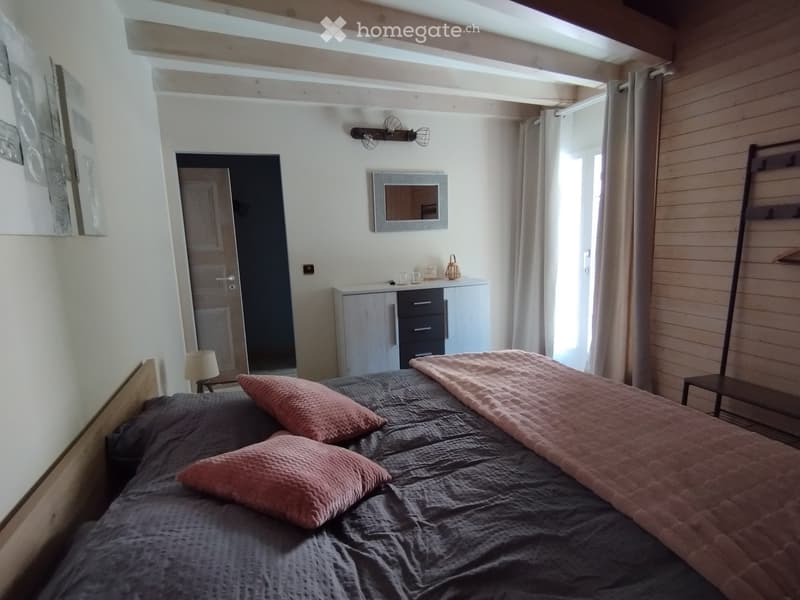Cozy bedroom with a wooden bed, grey bedding, pink pillows, a nightstand with a lamp, a cabinet with drawers, a mirror, and a wardrobe. The room features exposed wooden beams and large windows with curtains.