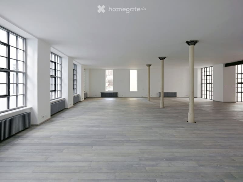 Empty room with wooden flooring, white walls, large windows, and industrial-style columns.