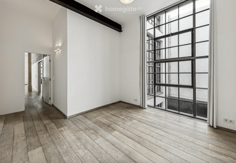 Empty room with light wooden flooring, white walls, large industrial-style window, and a door leading to another space.