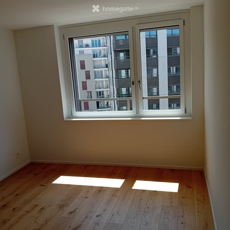 Empty room with wooden flooring and large windows showing a view of neighboring buildings.