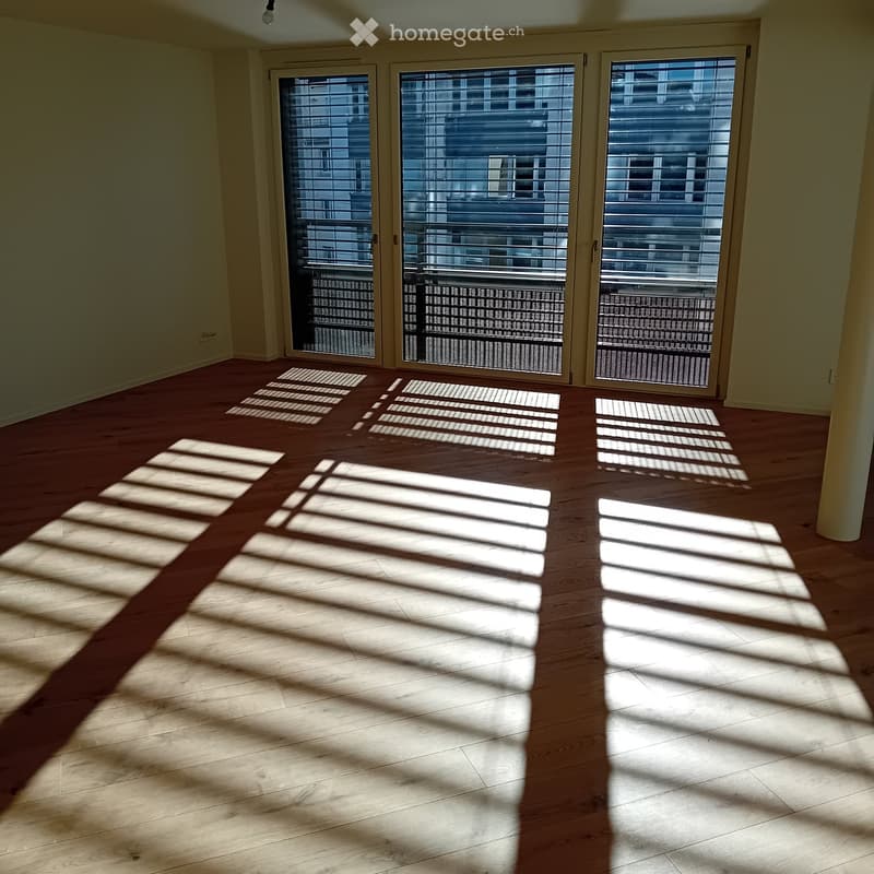 Empty room with wooden flooring and large sliding glass doors leading to a balcony. Sunlight streams in, casting shadows on the floor.