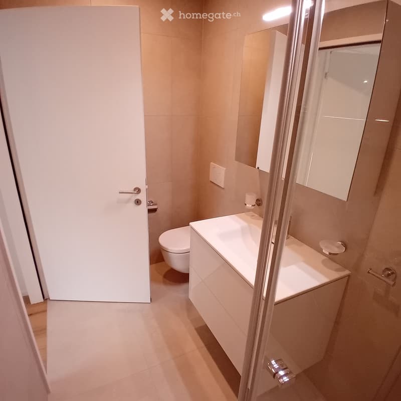 Modern bathroom with a white toilet, sink, and mirrored cabinet. The space features light-colored tiles and a glass shower enclosure.