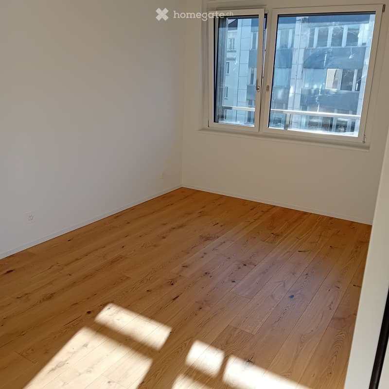 Empty room with light wooden flooring and white walls, featuring a large window with a view of neighboring buildings.