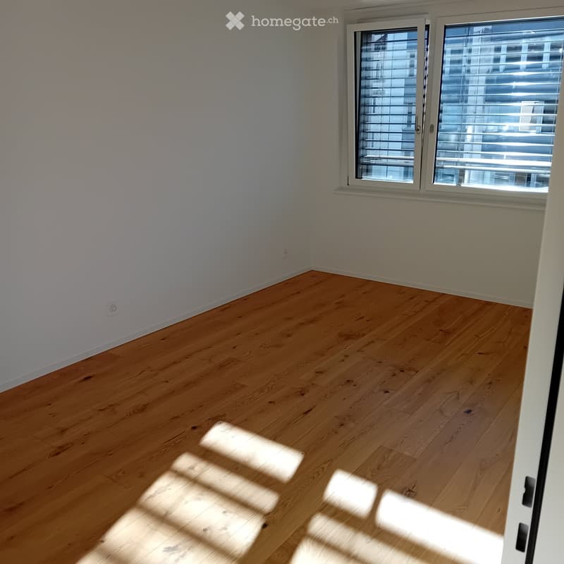 Empty room with light wooden flooring and white walls, featuring a large window with blinds allowing natural light to enter.