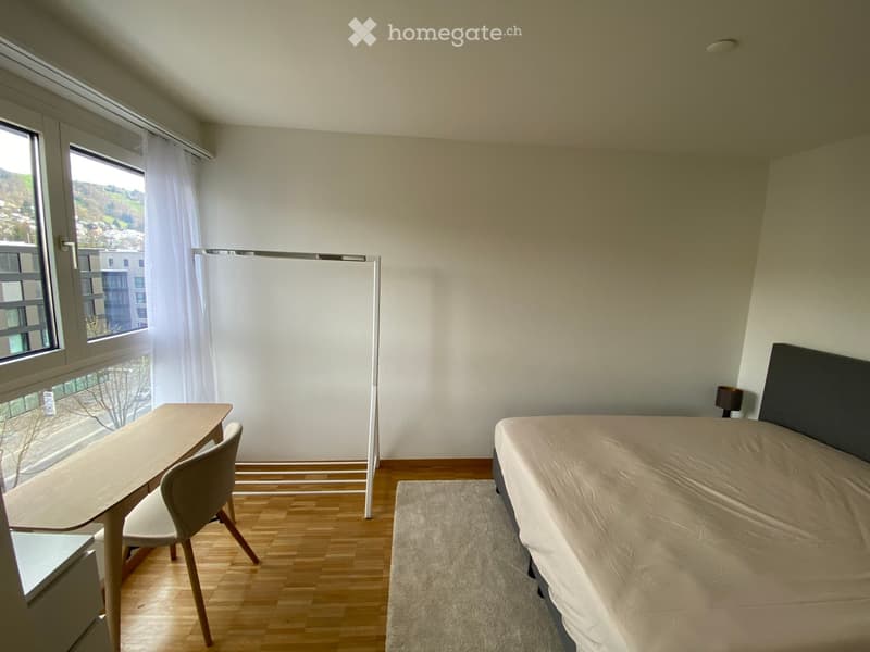 Modern, minimalist bedroom with a bed, a small table, and a chair. The room features wooden flooring, large windows with white curtains, and a simple, contemporary design.