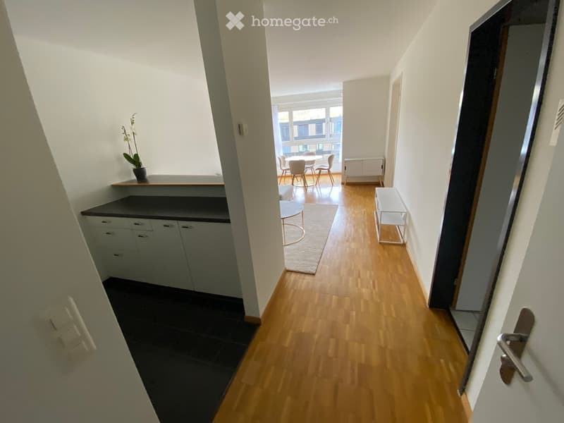 A modern hallway with wooden flooring, white walls, and a small kitchen area visible on the left. A dining area with chairs and a table is visible in the background, along with a small round table and a rug.