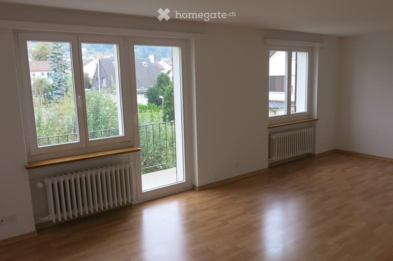 Empty room with wooden flooring, large windows, radiators, and a sliding glass door leading to a balcony.