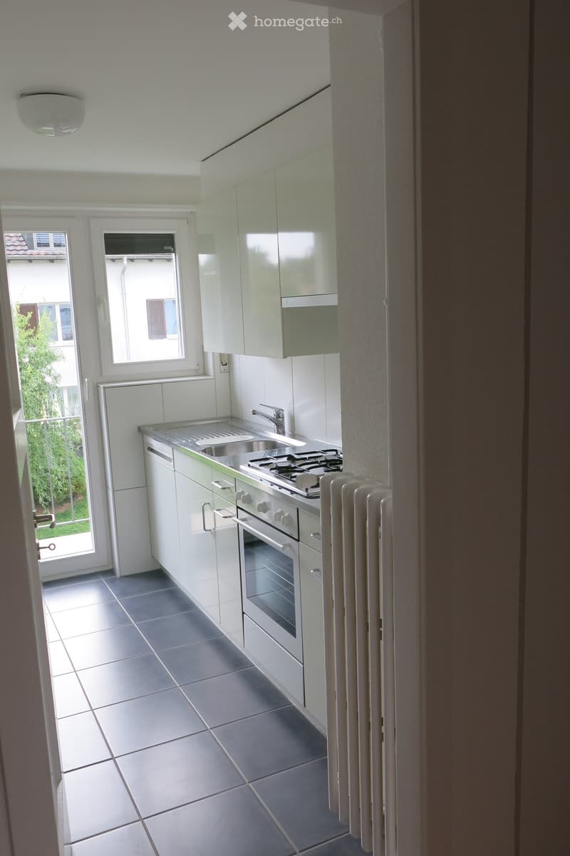 Modern kitchen with white cabinets, stainless steel sink, stove, and oven. Grey tiled floor and white walls.