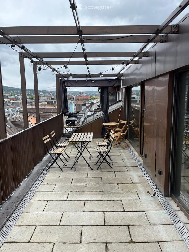 Roof terrace with wooden tables and chairs, overlooking a cityscape with buildings and mountains.