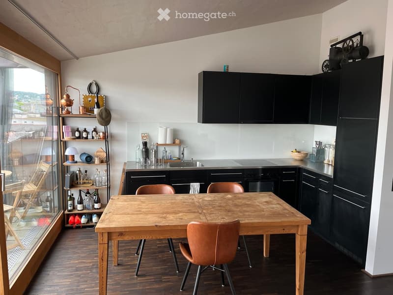 Modern kitchen with dark cabinets, wooden dining table, and leather chairs. Shelves hold various bottles and decorative items. Large window offers a view of the city.
