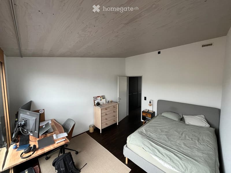 Modern bedroom with a bed, desk, and various office equipment. The room features a wooden floor, white walls, and a slanted wooden ceiling.