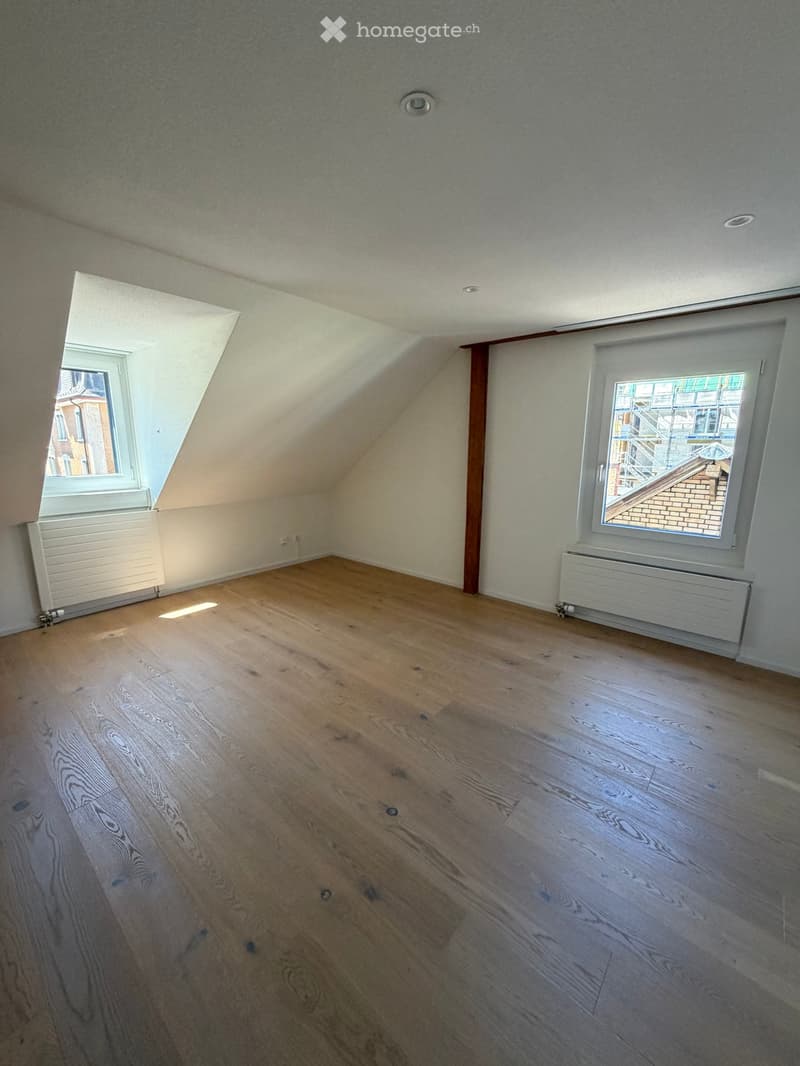 Empty attic room with wooden flooring, white walls, and two windows offering natural light. Radiators are present along the walls.