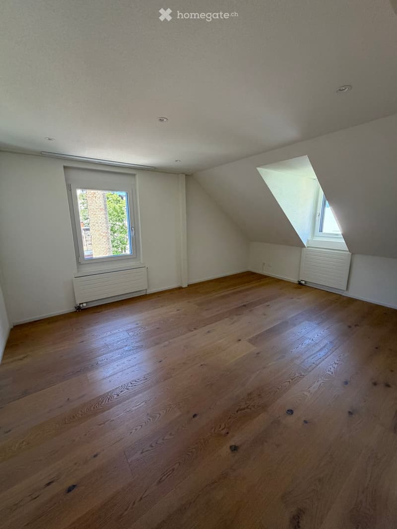 Empty attic room with wooden flooring, white walls, and two windows offering natural light. Radiators are visible under the windows.