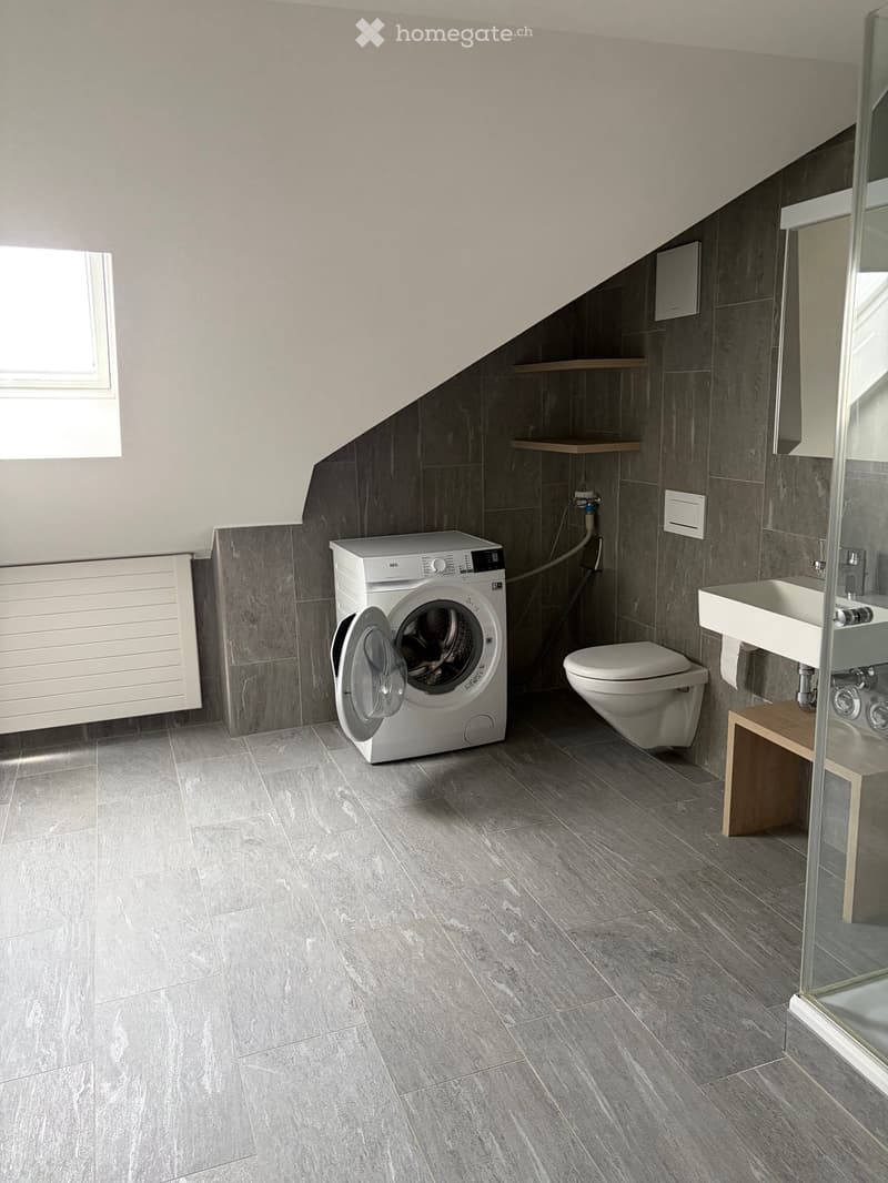 Modern bathroom with grey tiled walls and floor, white washing machine, toilet, sink, and small wooden stool.