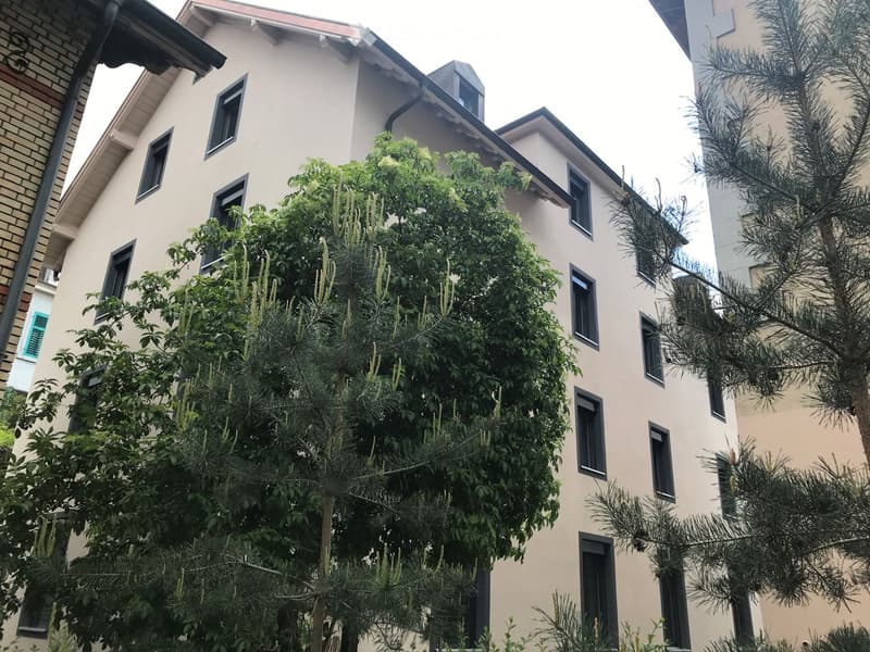Multi-story residential building with light beige facade, multiple windows, and surrounding greenery including trees and shrubs.