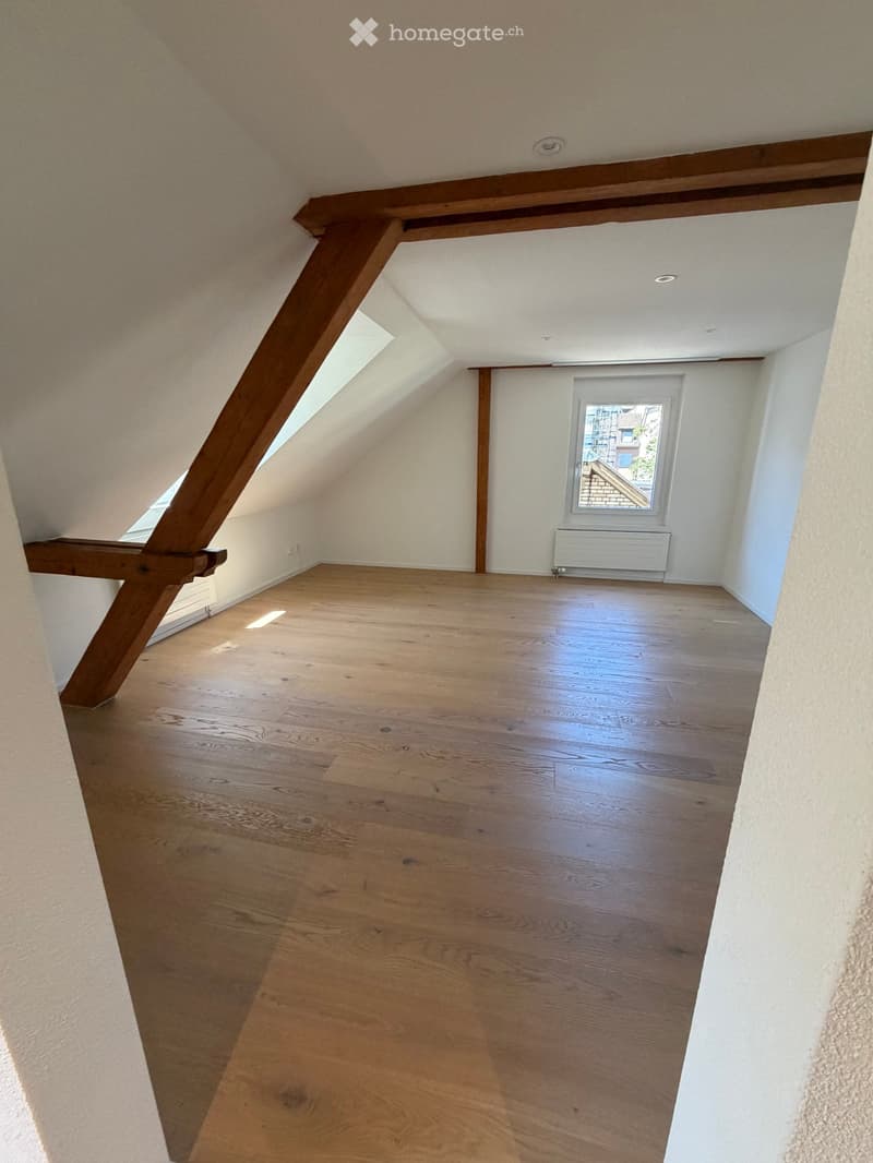 Empty attic room with wooden flooring, white walls, exposed wooden beams, skylight windows, and a radiator.