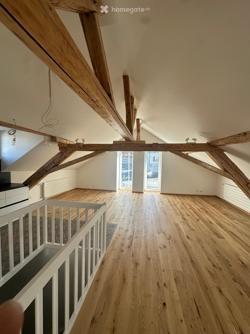 Empty attic space with wooden beams, wooden flooring, and large windows offering a view of the street.