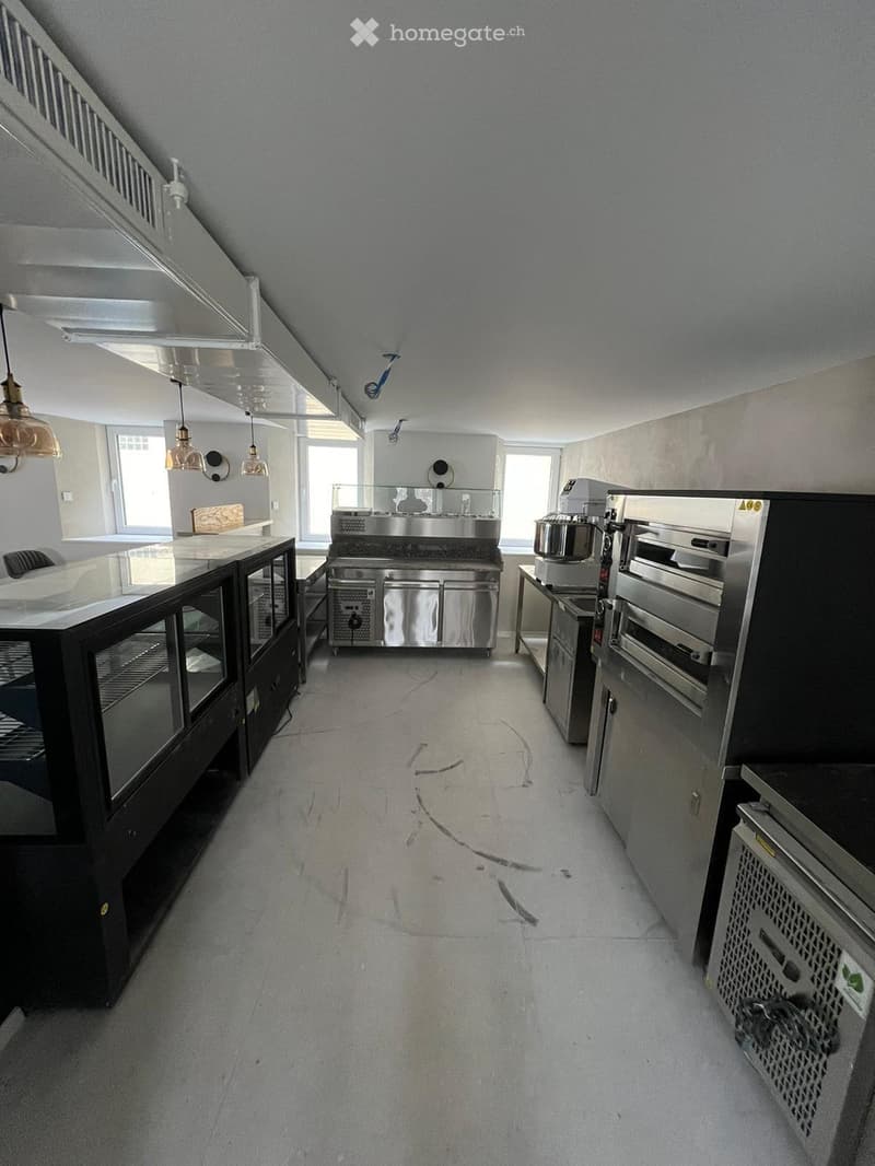 Commercial kitchen with stainless steel appliances, including a refrigerator, oven, and sink, along with a counter and pantry. The space features pendant lighting and large windows.