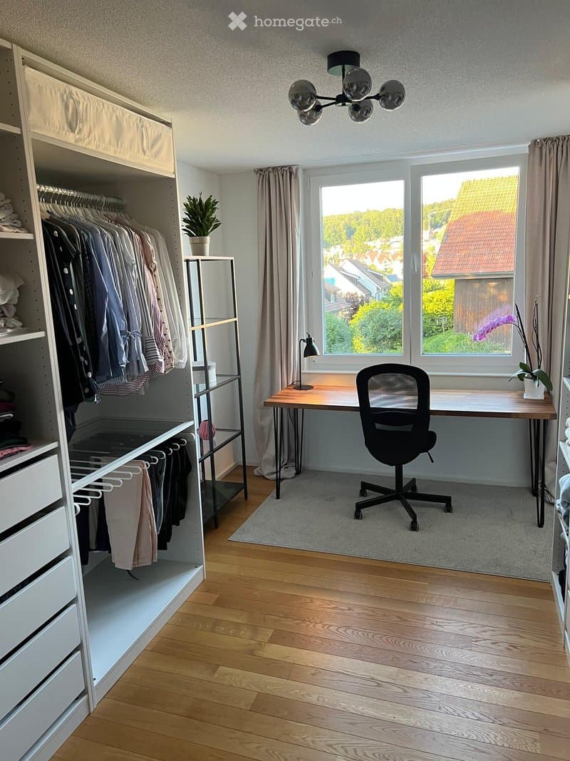 Modern, well-lit room with wooden flooring, large windows offering a view, a desk with a chair, a wardrobe, and decorative plants.