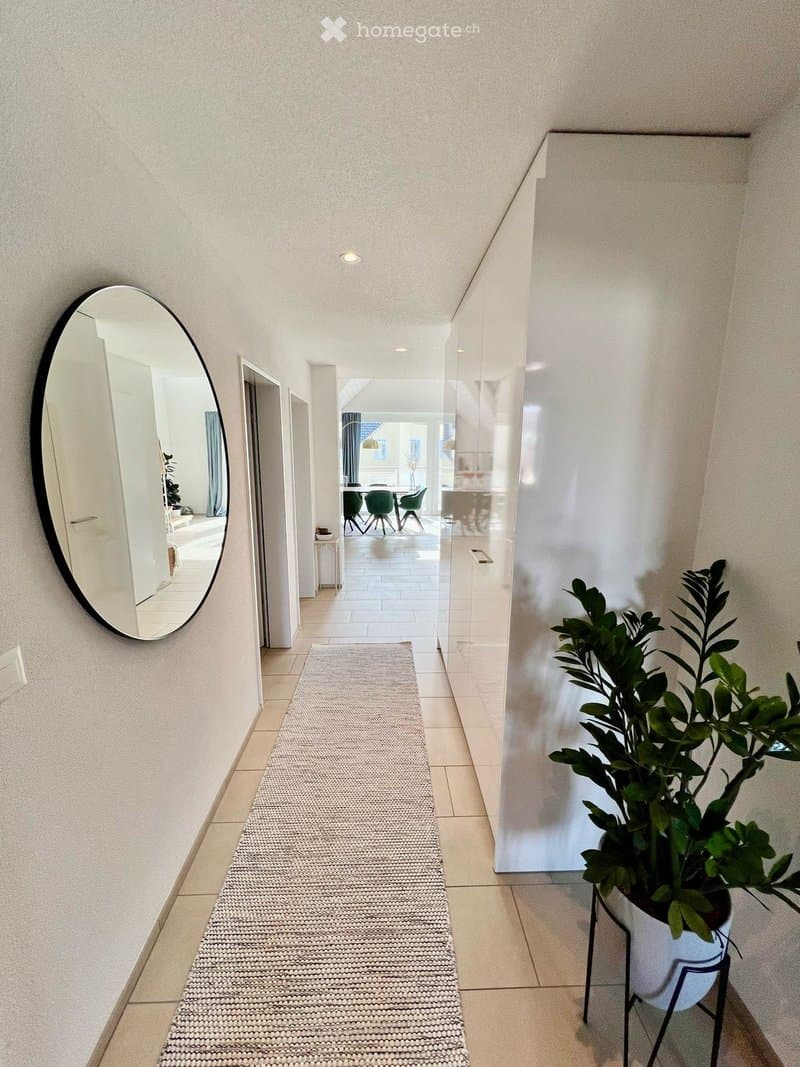 Modern hallway with tiled flooring, a round wall mirror, a potted plant, and a textured runner. The hallway leads to an open area with visible furniture.