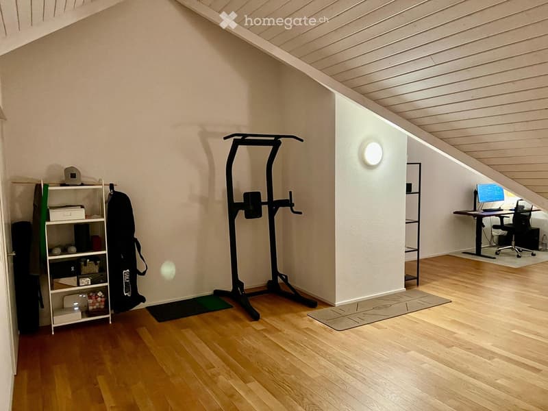 A multi-purpose room with wooden flooring, a shelf with storage items, a fitness machine, a yoga mat, and a desk with a laptop and chair.