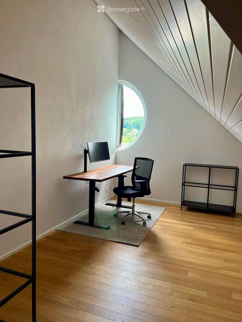 Modern, minimalist room with wooden flooring, a desk, ergonomic chair, and a large round window offering a scenic view.