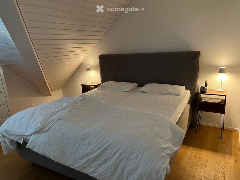 Cozy bedroom with a large bed, nightstands, lamps, and an alarm clock. The room features wooden flooring and a slanted ceiling with a skylight.