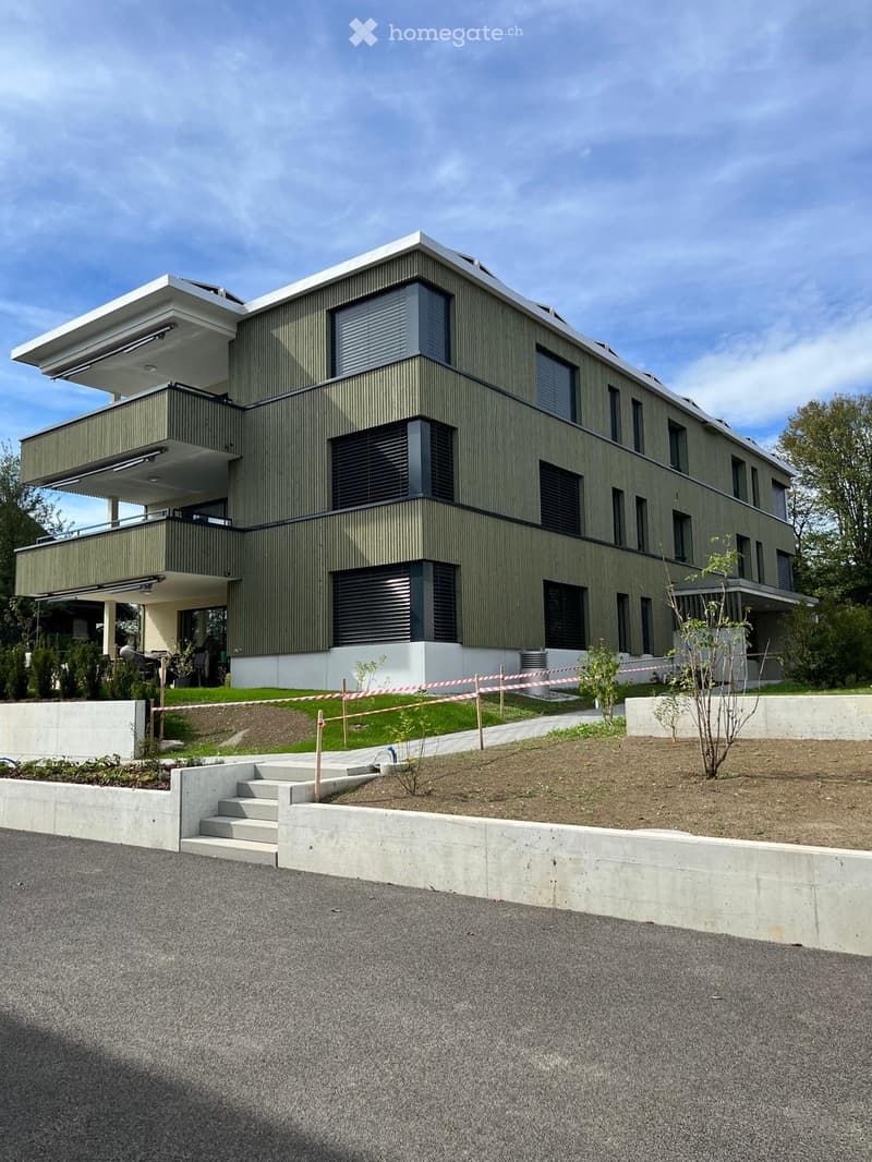 Modern two-story house with green siding, large windows, and balconies. The house features a raised ground floor and is surrounded by a landscaped garden area.