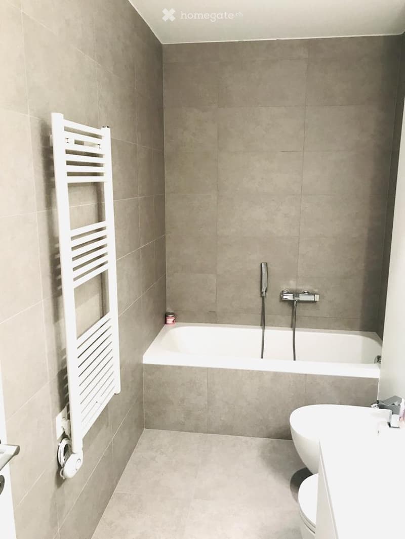 Modern bathroom with beige tiles, a bathtub, toilet, sink, and a white radiator on the wall.