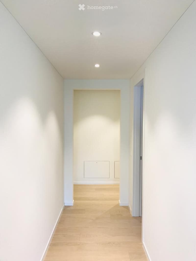 A clean, modern hallway with light wooden flooring, white walls, and recessed ceiling lighting. The hallway features a single door at the end and a partially open door on the right side.