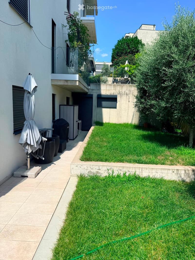 Outdoor patio area with a tiled walkway, green lawn, and outdoor furniture including a closed umbrella, chairs, and a small table.