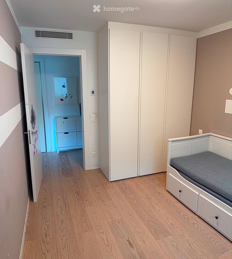 Bedroom with a single bed, white wardrobe, and wooden flooring. An open doorway leads to a small room with a refrigerator.