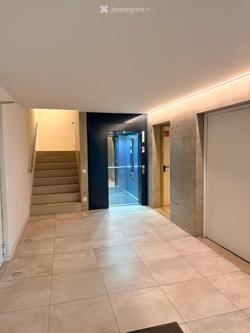 Modern hallway with tiled floor, staircase to the left, elevator in the center, and a door to the right.